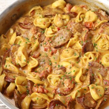 Saute pan filled with tortellini and sausage on a white wood counter.