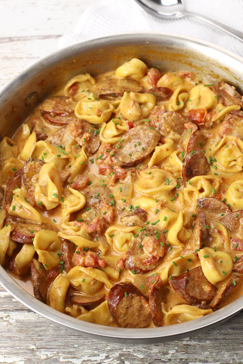 Saute pan filled with tortellini and sausage on a white wood counter.