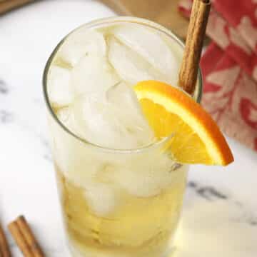 A highball glass filled with a pale orange cocktail garnished with an orange slice and cinnamon stick.