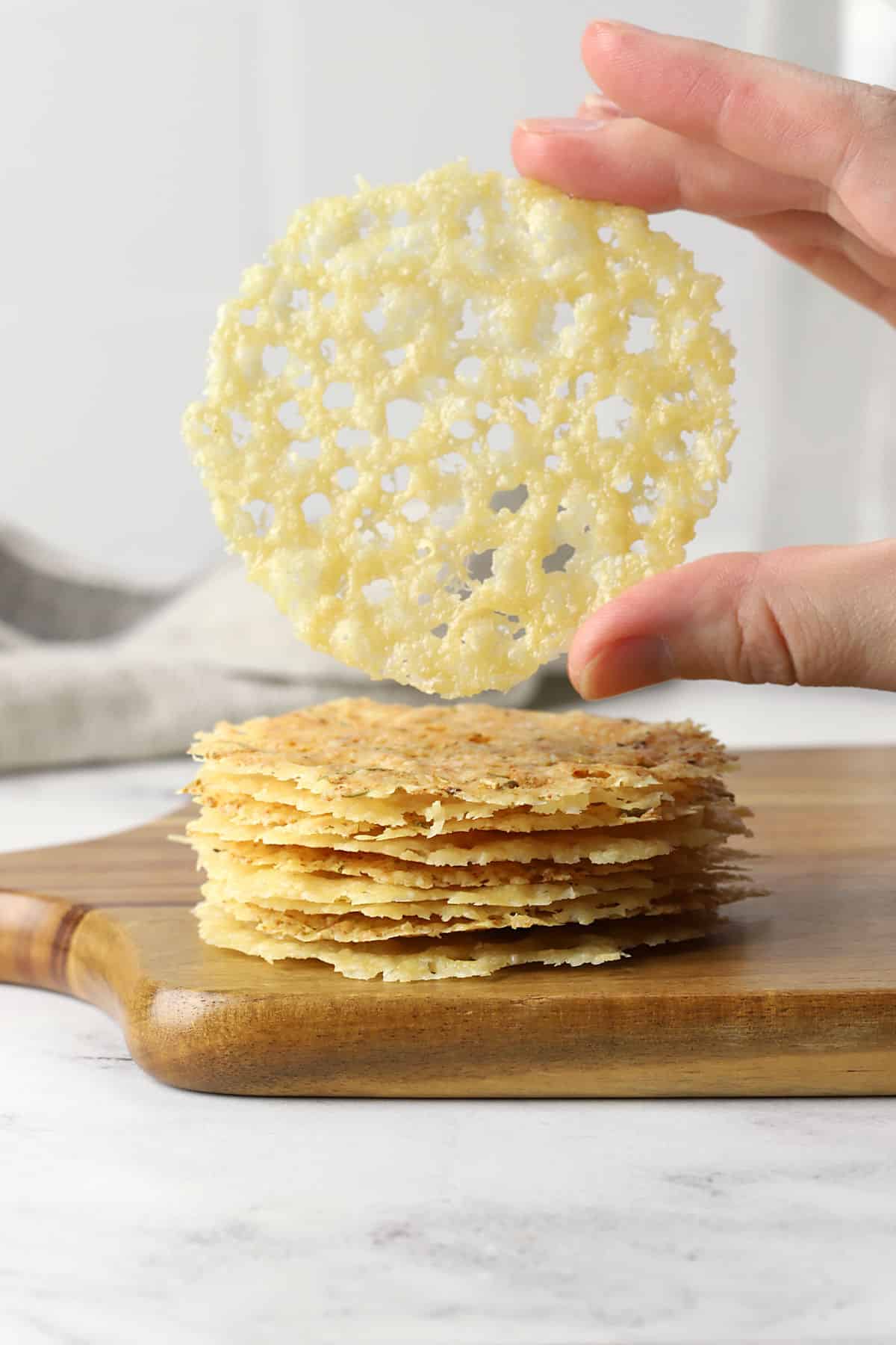 A hand holding a single parmesan crisp above a stack of more parmesan crisps.