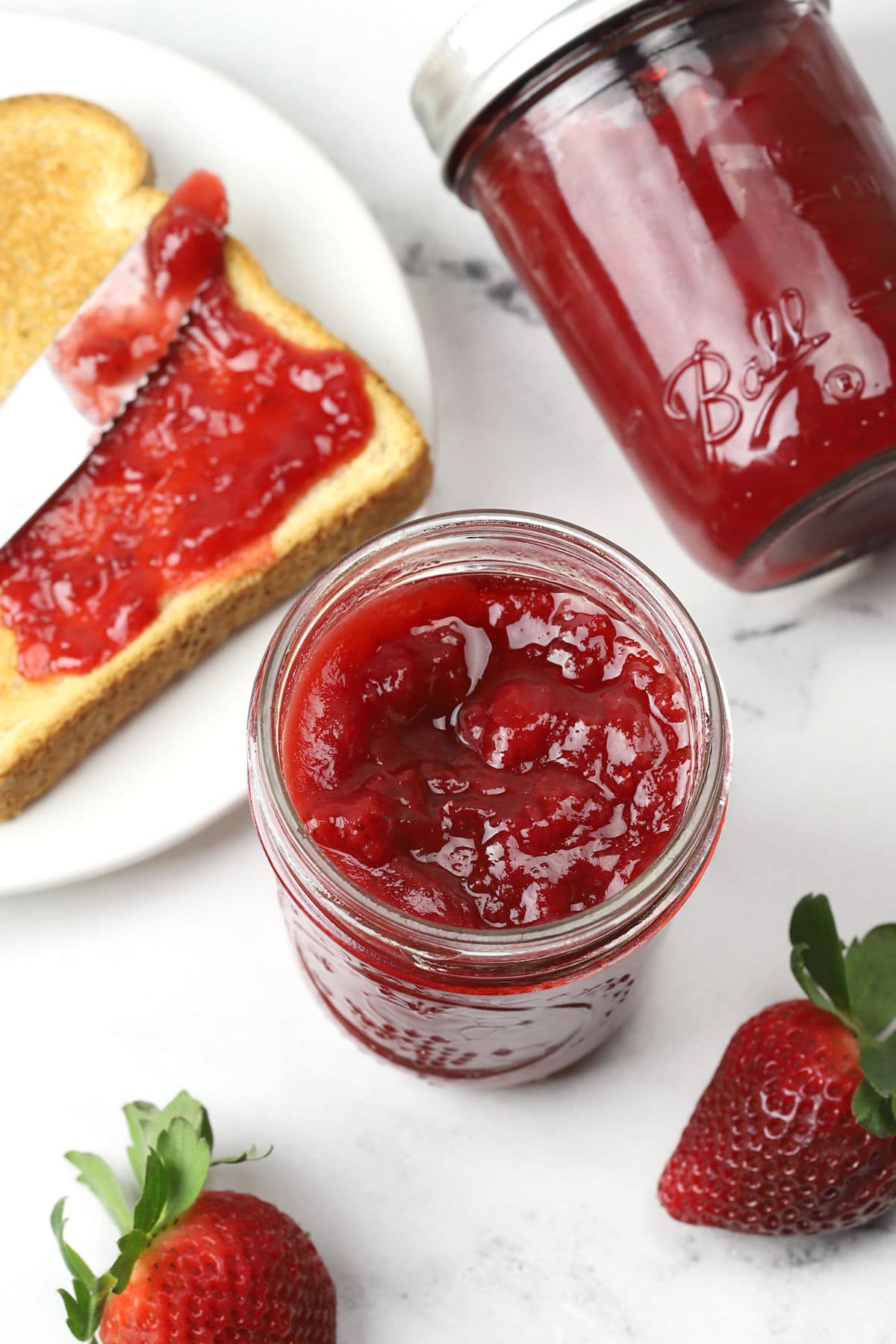 A jar of opened strawberry jam with a piece of toast on a plate.