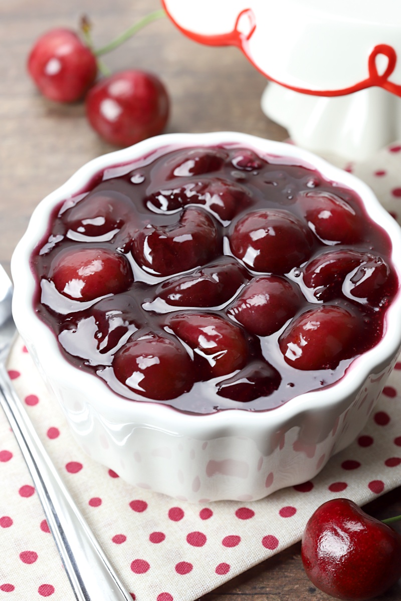 Pie filling in a bowl, a spoon, and a few cherries on a counter top. 