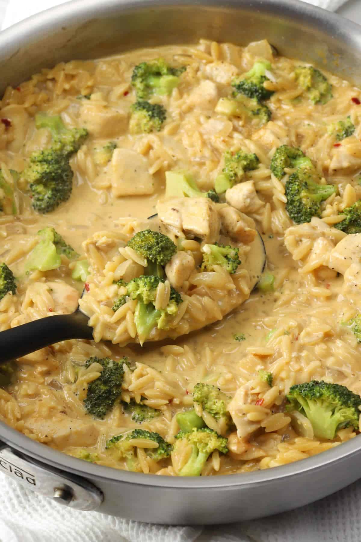 A serving spoon scooping cheesy chicken broccoli orzo from a sauté pan.