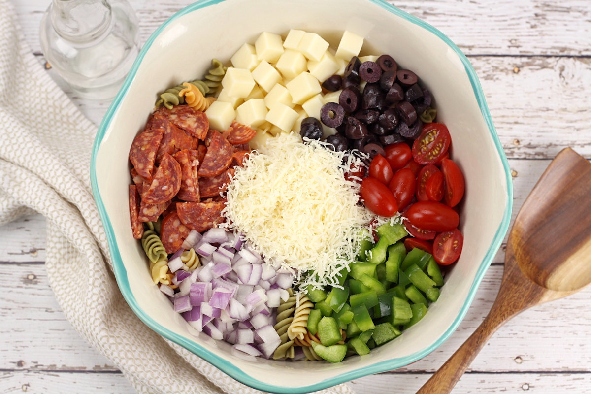 Ingredients for a classic pasta salad in a teal bowl, before being mixed together - mozzarella cheese, olives, cherry tomatoes, green bell pepper, red onion, pepperoni, and parmesan cheese.