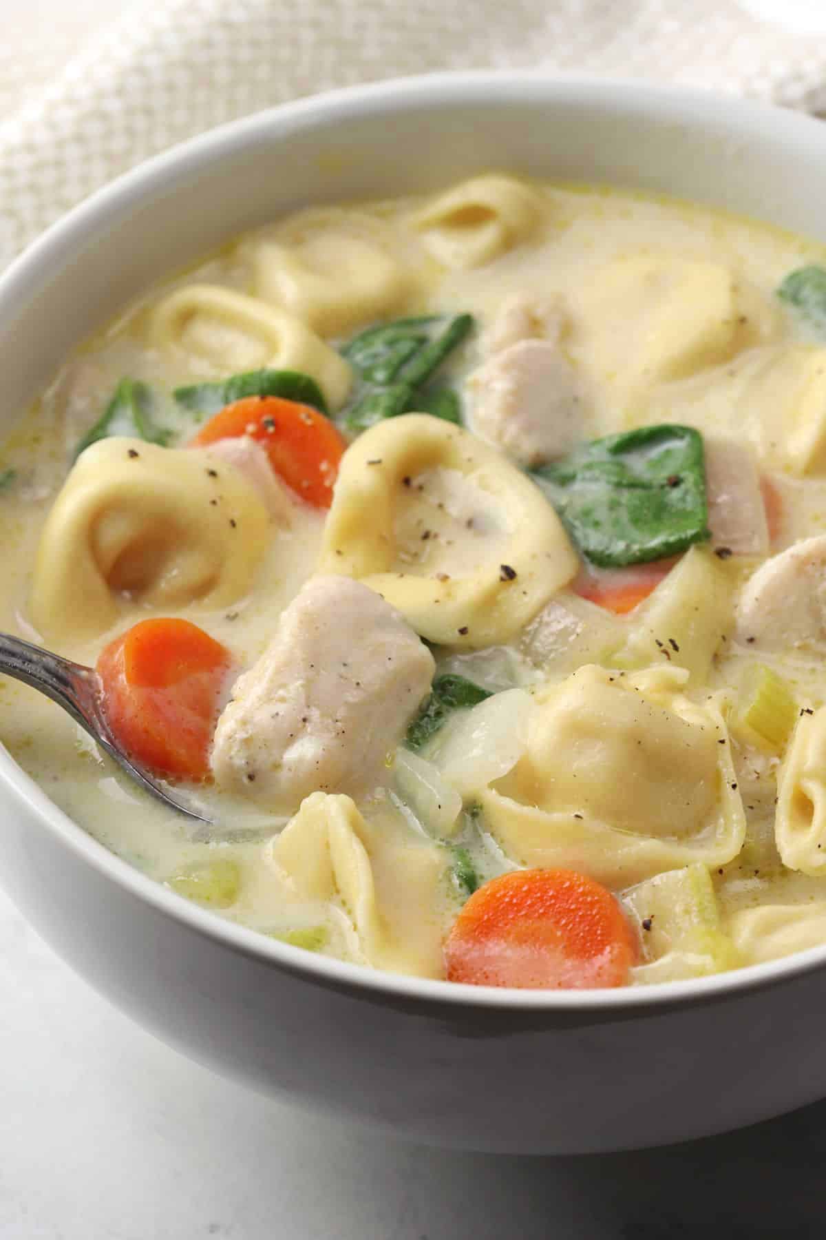 A metal spoon scooping tortellini soup from a white bowl.