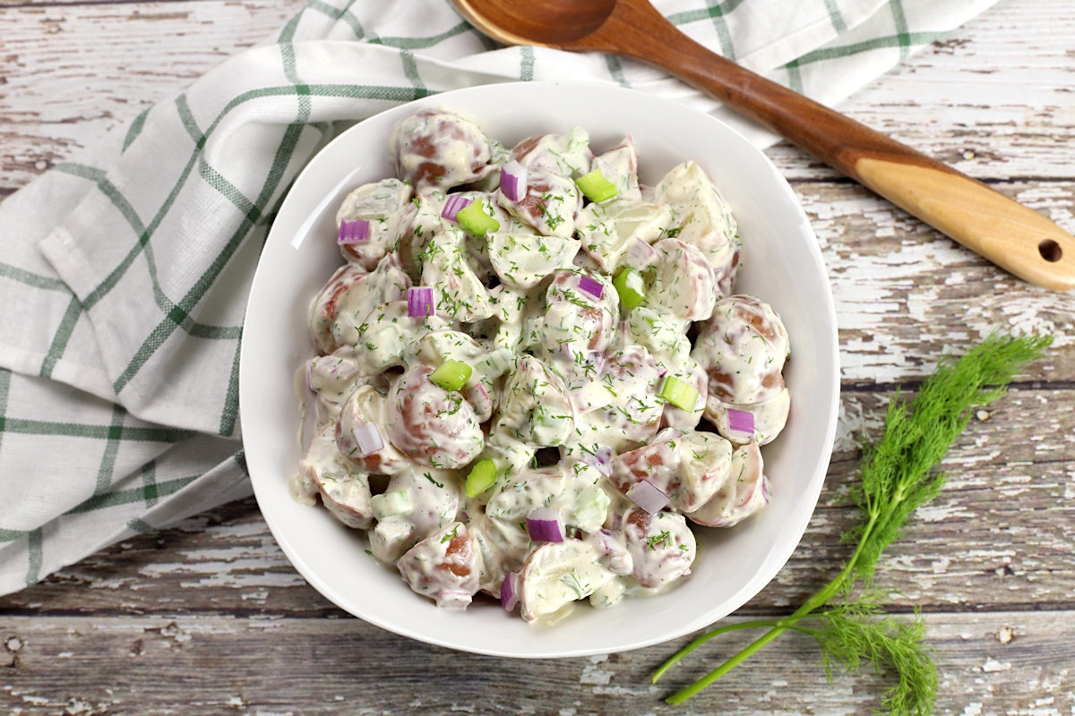 White bowl filled with potato salad, with a kitchen towel and wooden spoon.