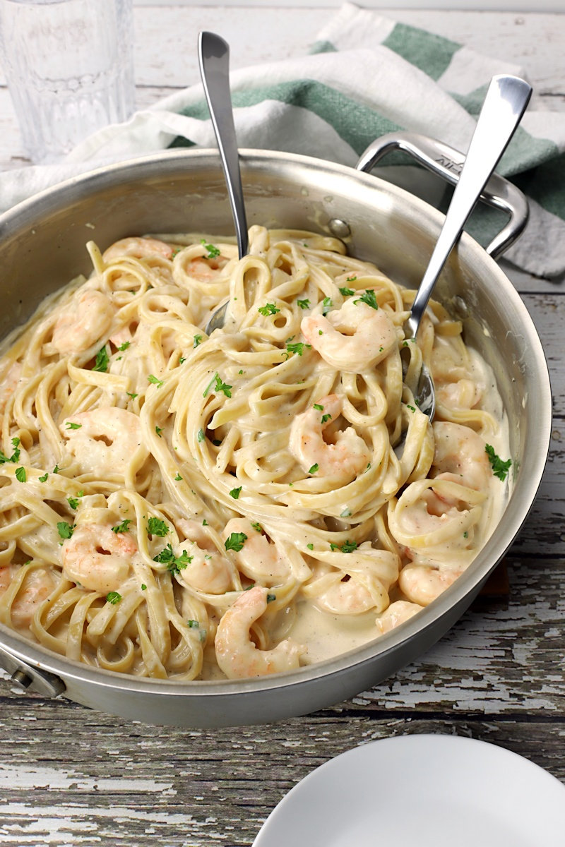 Metal fork and spoon servers scooping shrimp fettuccine alfredo out of pan.