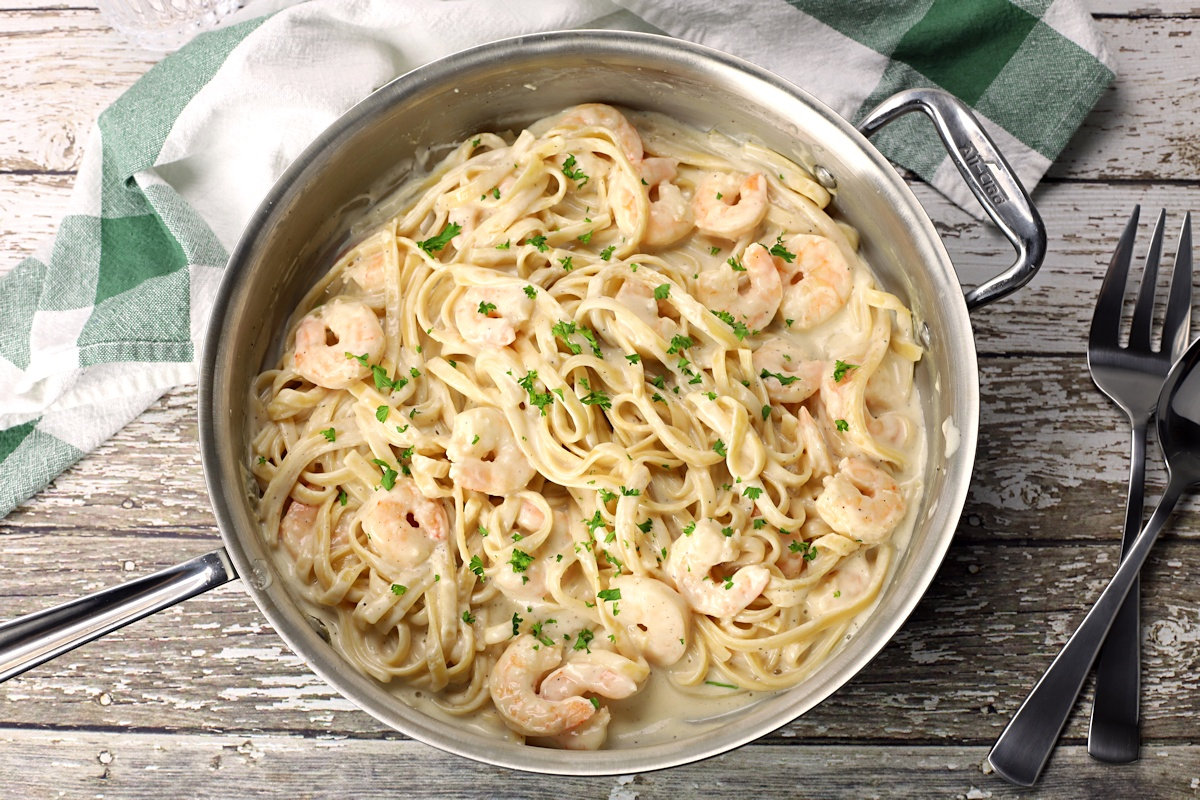 Saute pan filled with fettuccine noodles and shrimp on a wood countertop.