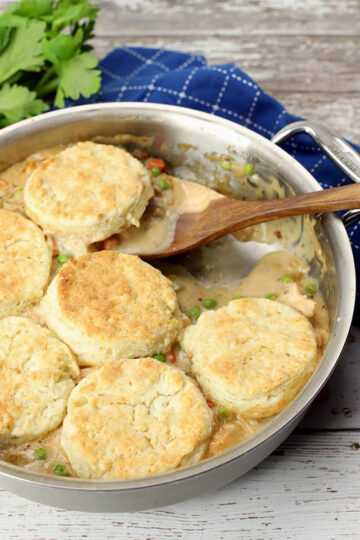 Close up of a wooden serving spoon scooping chicken pot pie topped with biscuits from a sauté pan.