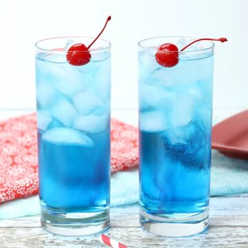 Two blue cocktails on a white wood countertop with cherries and straws.
