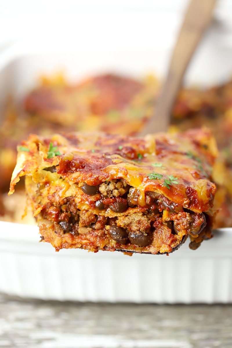 A serving spoon scooping beef enchilada casserole from a serving dish.