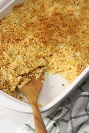 A wooden serving spoon scooping hash brown potato casserole from a white casserole dish.