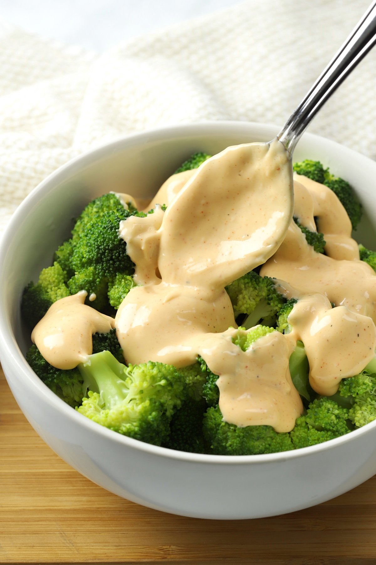 A metal spoon pouring cheese sauce onto a bowl of broccoli.