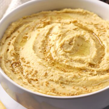A white bowl filled with hummus, topped with sesame seeds and olive oil.