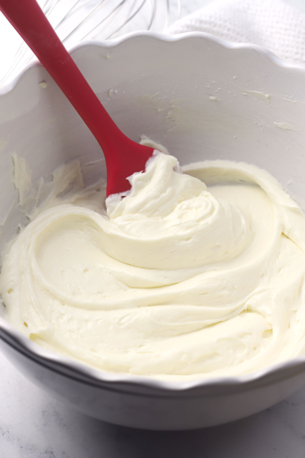 A white bowl filled with cream cheese frosting and a red spatula.