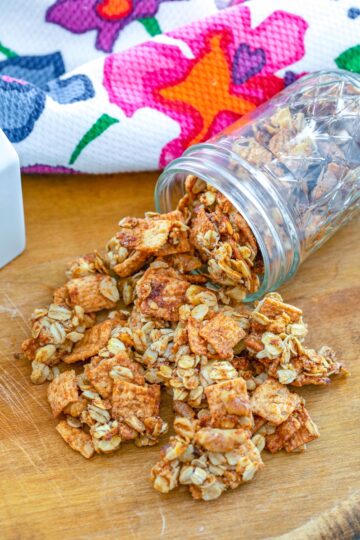 A jar of granola spilling onto a wooden cutting board with a floral kitchen towel.