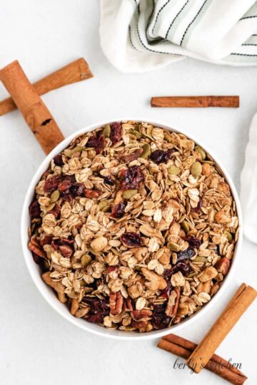 White bowl filled with granola, sitting with cinnamon sticks on a counter top.