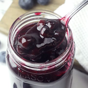 Metal spoon scooping blueberry topping from a glass jar.