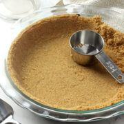 A metal measuring cup sitting in a partially prepared graham cracker crust in a glass pie pan.