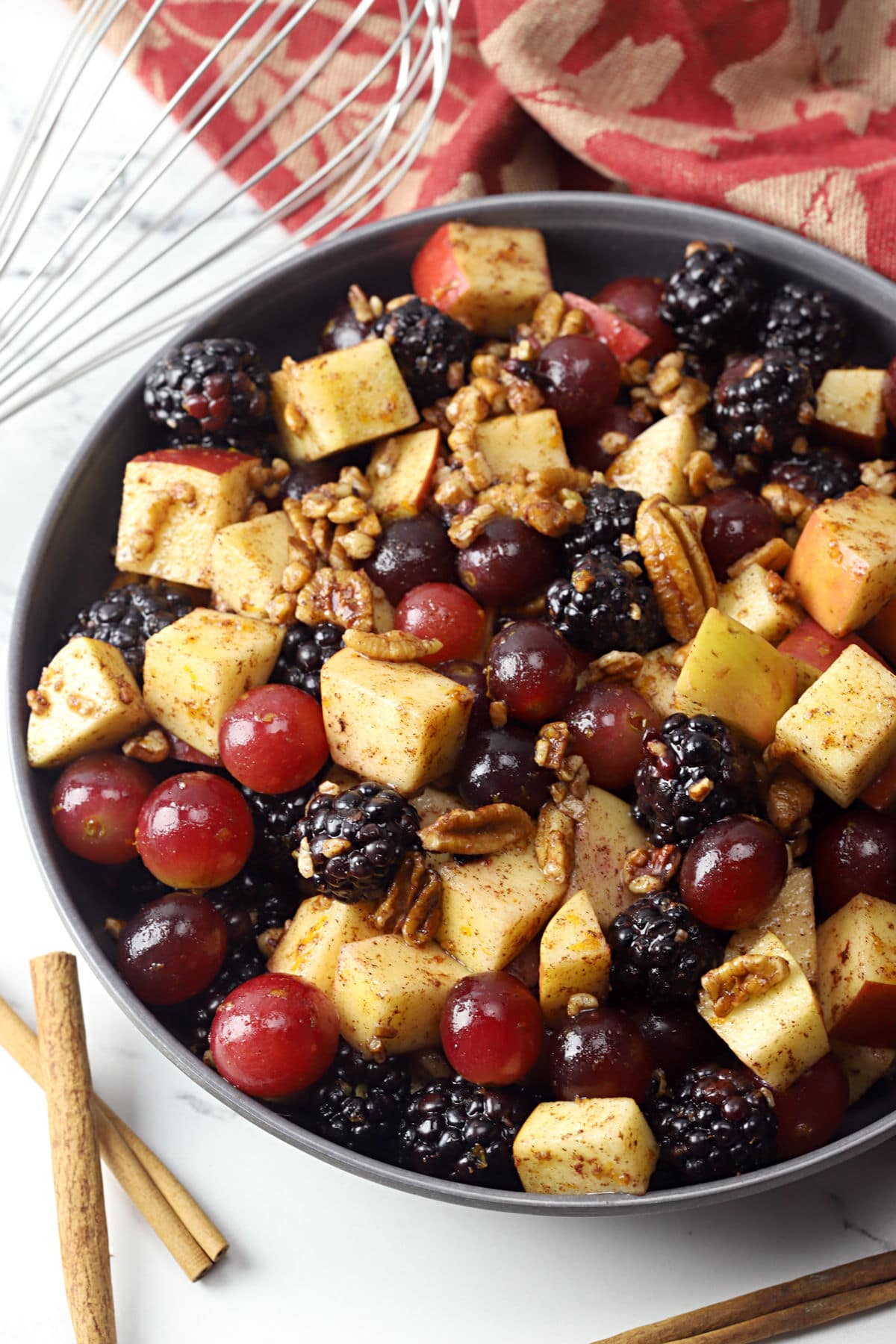 A bowl filled with fruit salad next a red and gold kitchen towel and cinnamon sticks.