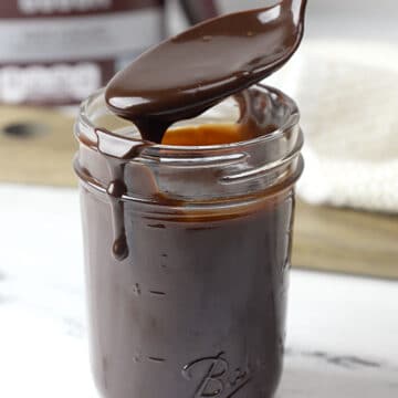 A metal spoon scooping hot fudge out of a glass jar.