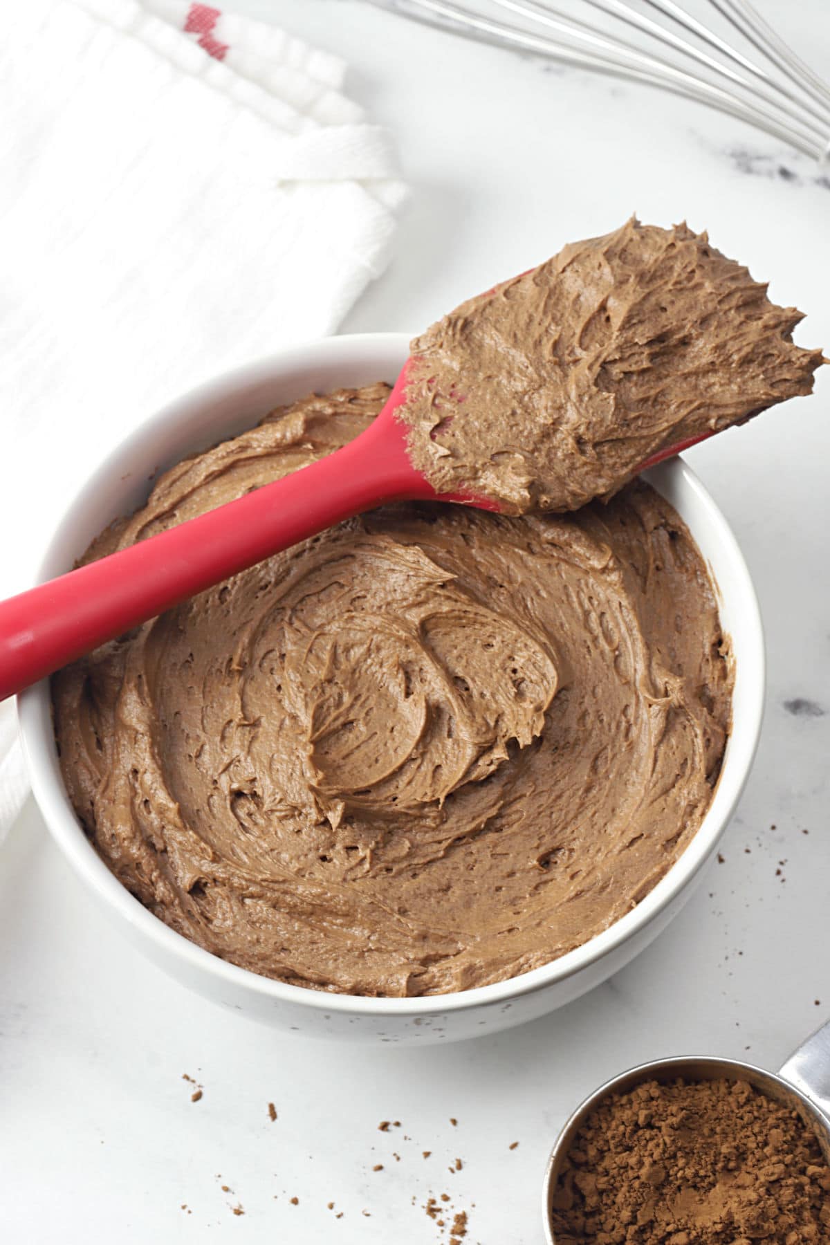 A white bowl filled with chocolate frosting, with a red spatula balanced across the top.