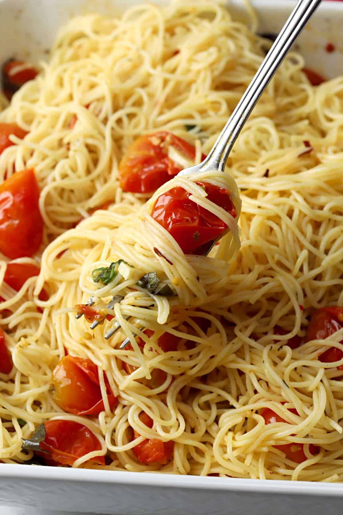 A fork filled with angel hair spaghetti, roasted tomatoes, and thin sliced garlic pieces.