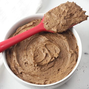 A white bowl filled with chocolate frosting, with a red spatula balanced across the top.