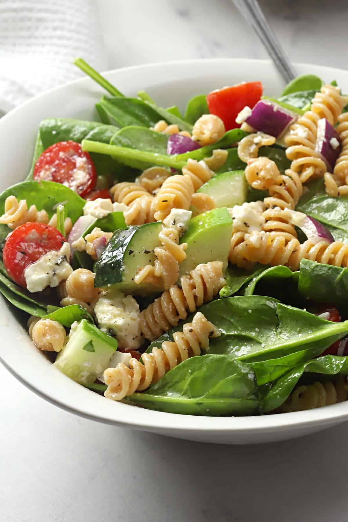 Pasta, cucumber, and tomatoes on top of a bed of spinach in a white bowl.