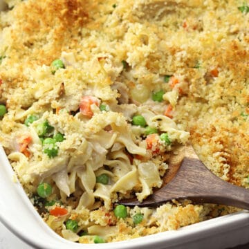 A wooden spoon scooping noodles, chicken, and veggies in a creamy sauce from a white casserole dish.