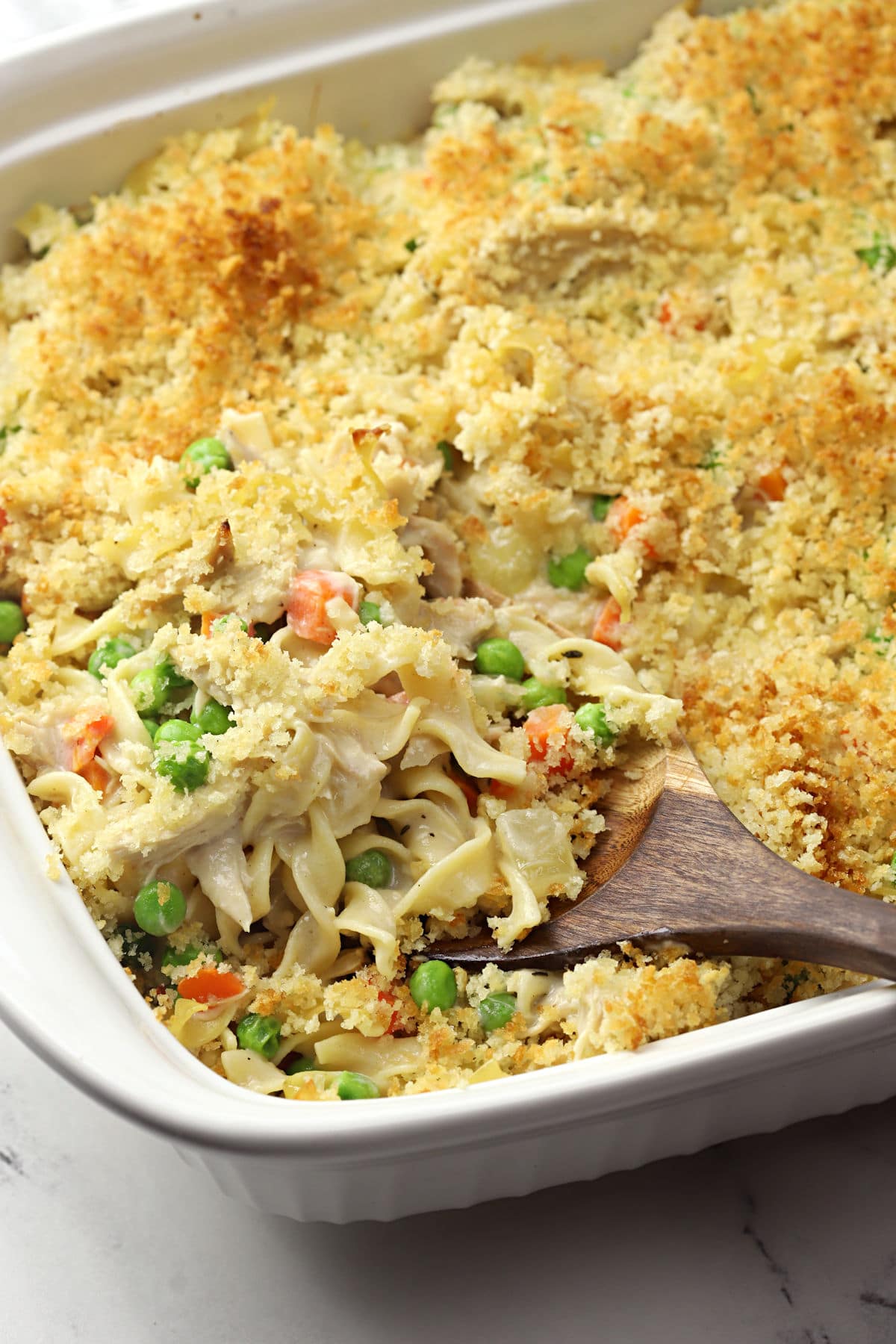A wooden spoon scooping noodles, chicken, and veggies in a creamy sauce from a white casserole dish.