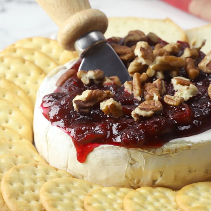 A whole wheel of brie cheese topped with cranberry sauce and pecans, with a serving knife and crackers.