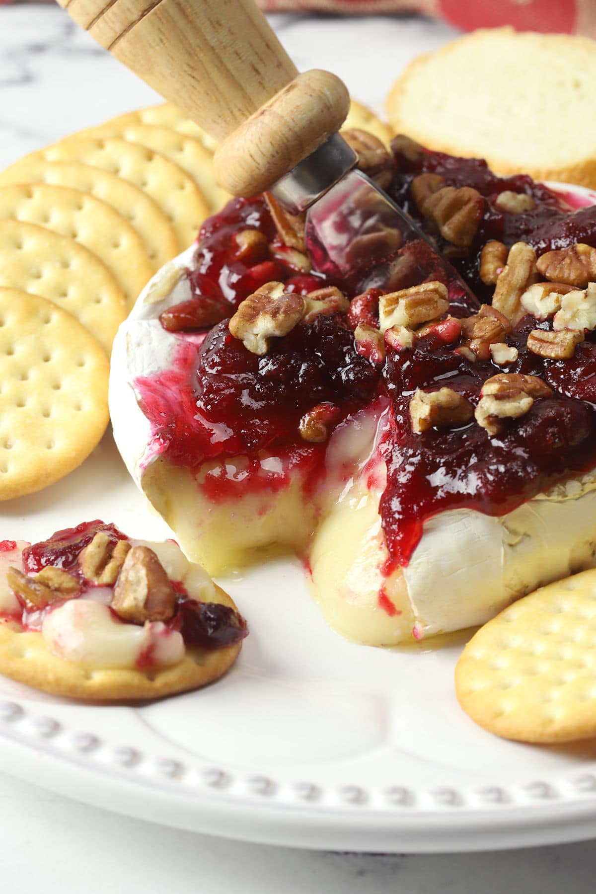 A wheel of melty brie cheese topped with cranberry sauce and pecans on a plate with crackers.