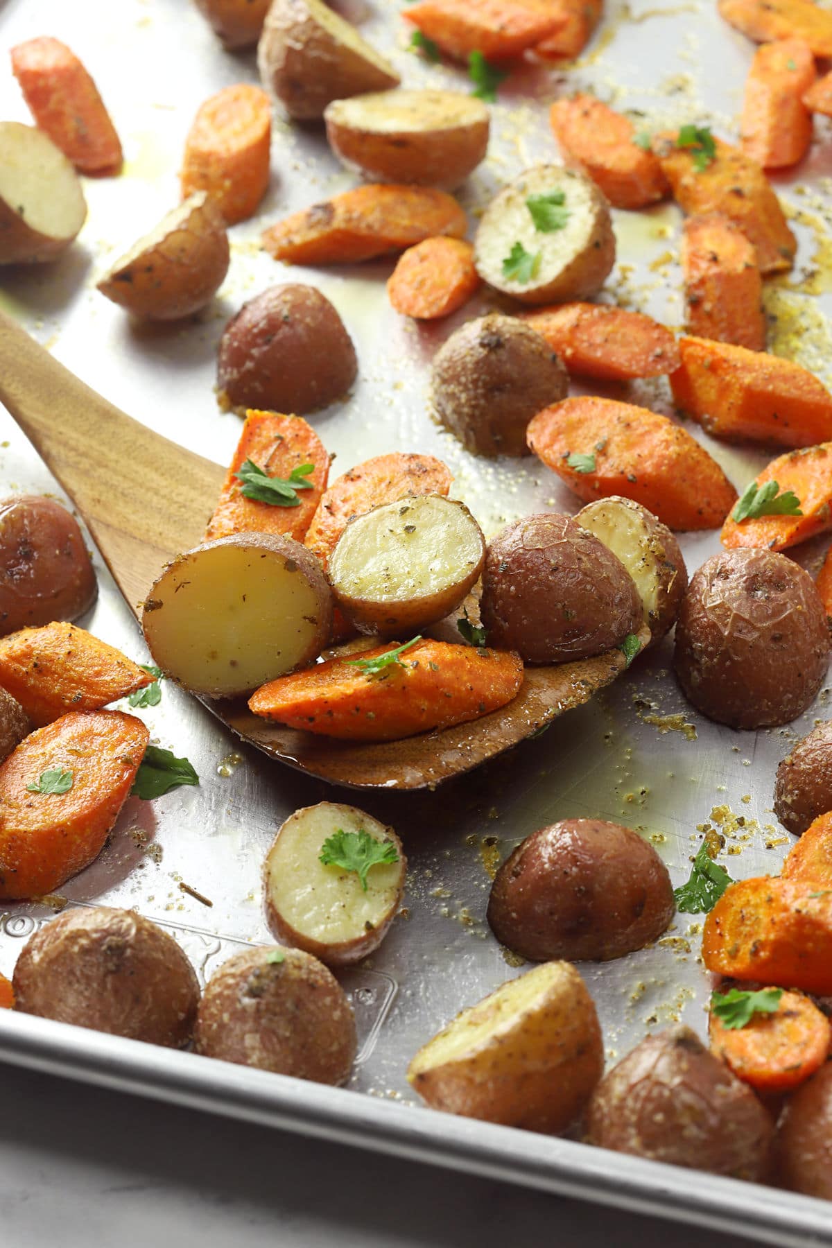 Wooden spatula scooping potatoes and carrots from a metal sheet pan.