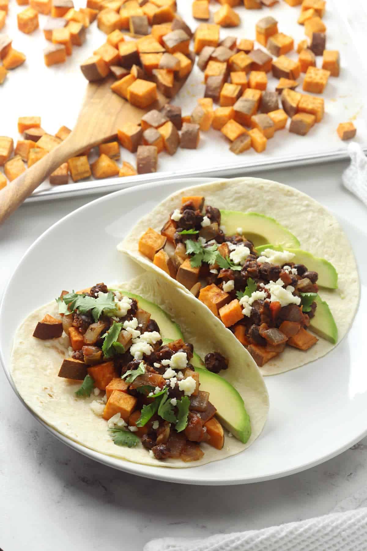 Two tacos on a white plate with a sheet pan of roasted sweet potatoes in the background.