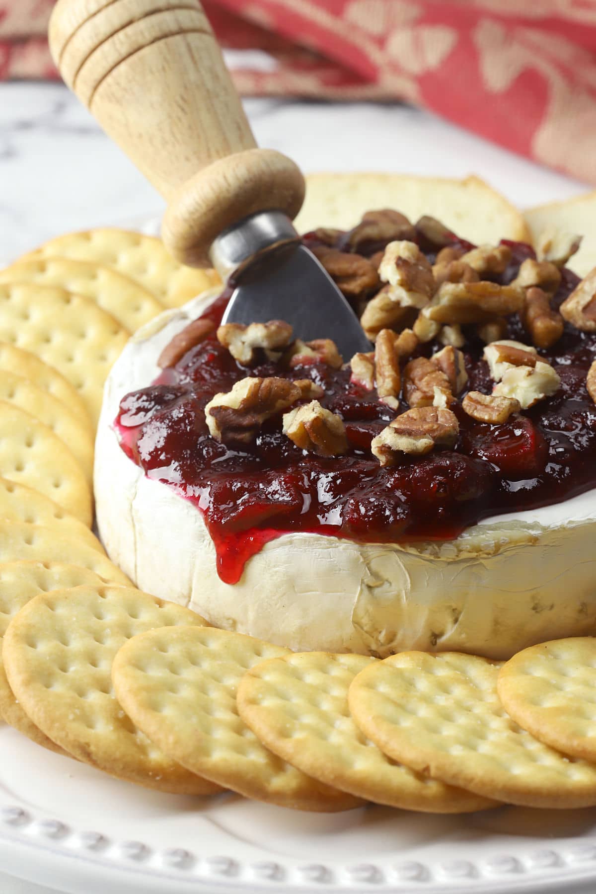 A whole wheel of brie cheese topped with cranberry sauce and pecans, with a serving knife and crackers.