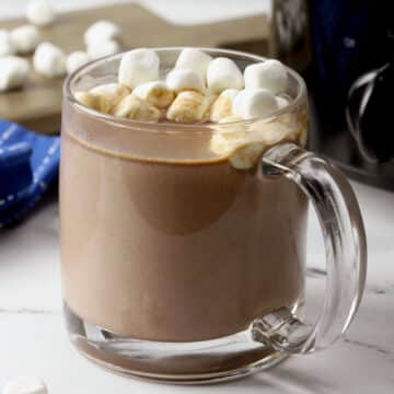 Glass mug of hot chocolate in front of a black slow cooker.