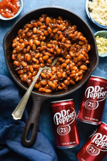 Baked beans in a cast iron skillet next to several cans of Dr. Pepper soda.