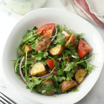 White bowl filled with arugula, potatoes, tomato, and red onion.