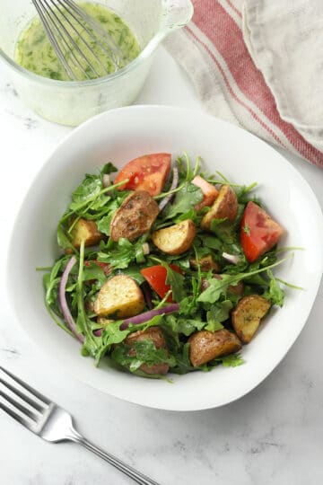 White bowl filled with arugula, potatoes, tomato, and red onion.