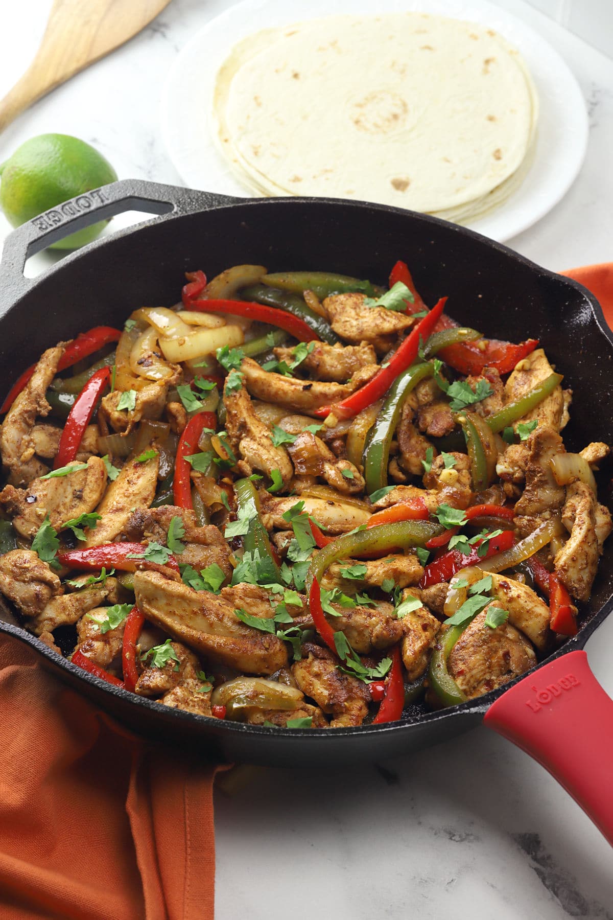 A cast iron skillet filled with cooked chicken fajitas, beside a plate of flour tortillas.