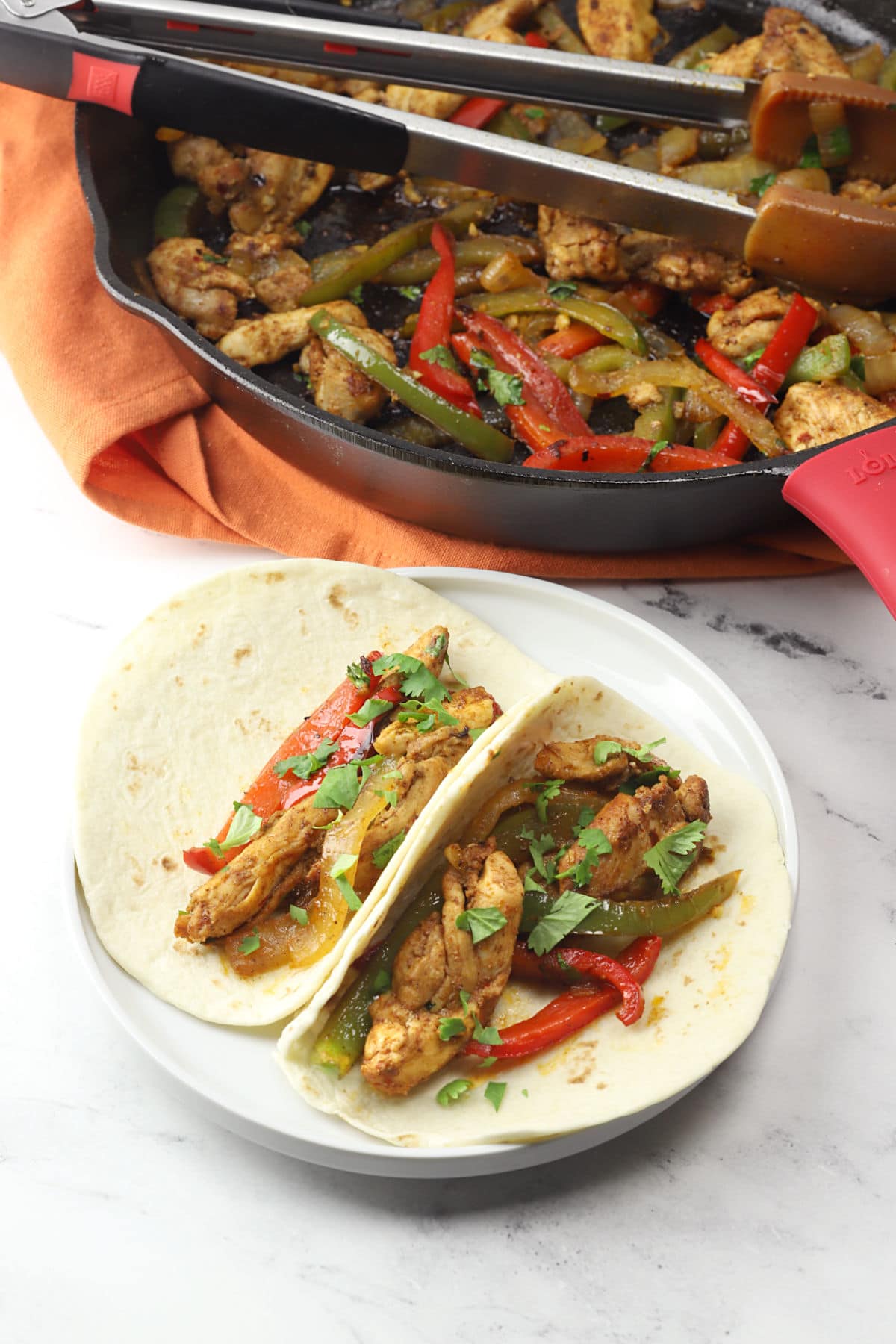Two flour tortillas filled with cooked chicken thighs, onions, and peppers, with a cast iron skillet of fajita filling in the background.