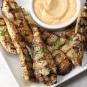A white serving plate of grilled chicken tenders with a bowl of creamy dipping sauce.