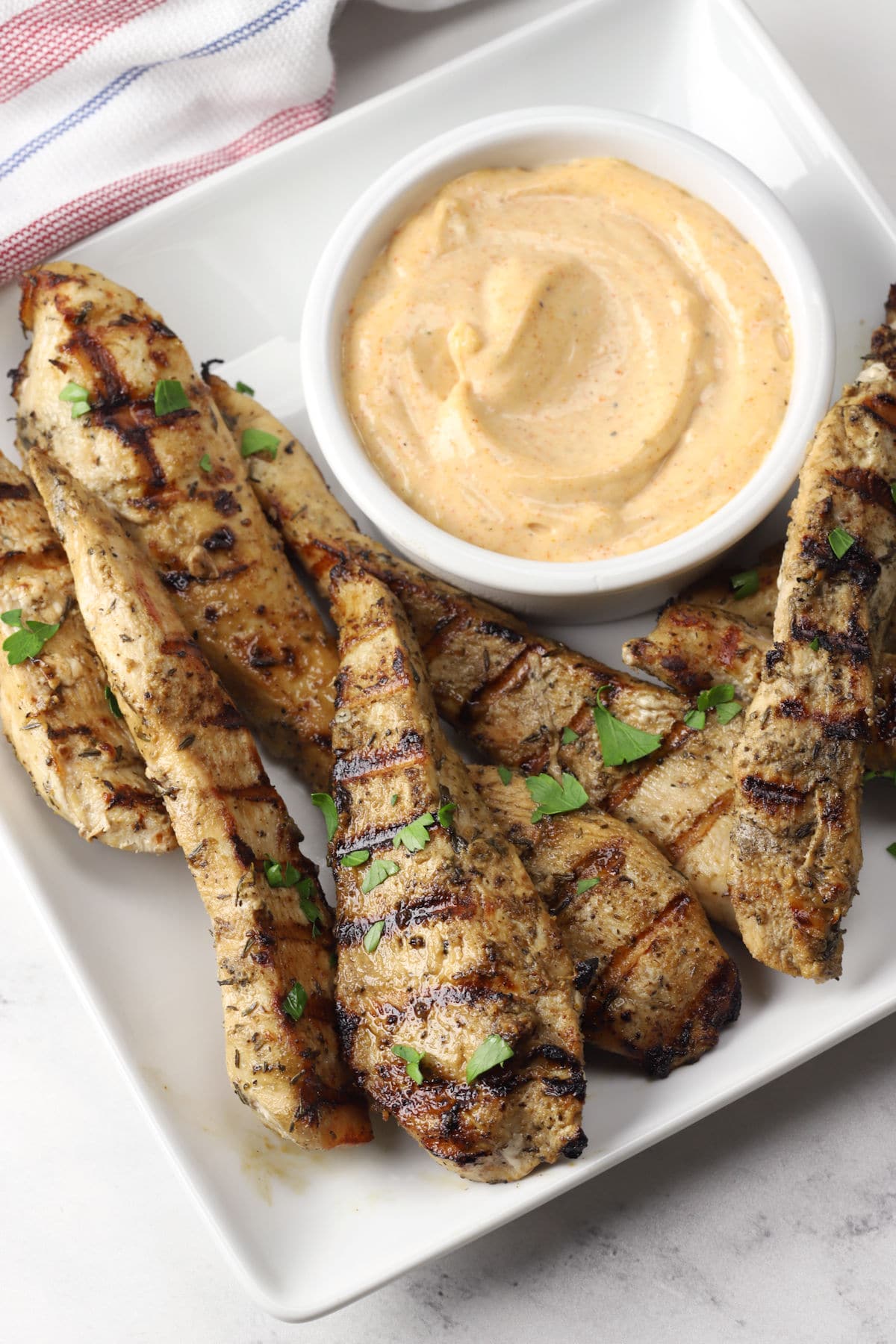 A white serving plate of grilled chicken tenders with a bowl of creamy dipping sauce.