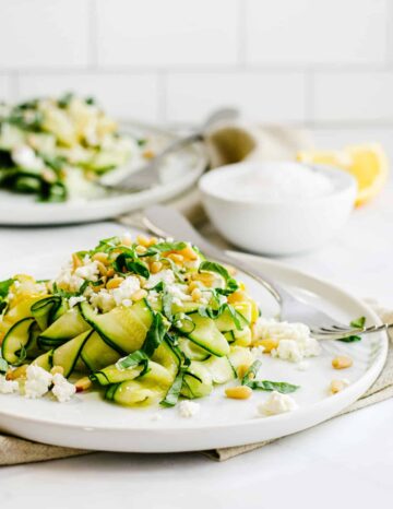 White plate filled with thin sliced zucchini salad with feta cheese and pine nuts.