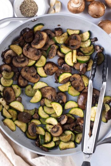Skillet filled with cooked mushrooms and zucchini, with a pair of tongs.