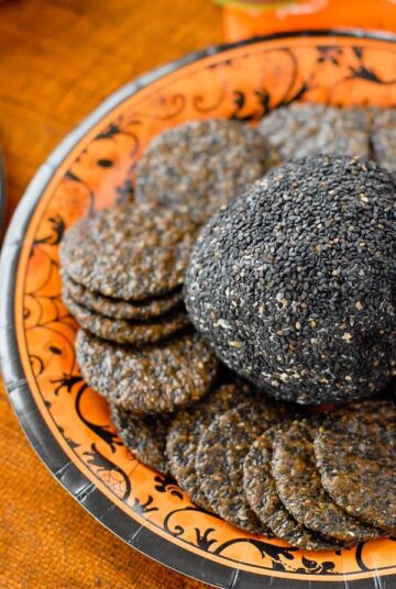 Cheese ball coated in black sesame seeds with crackers on an orange plate.