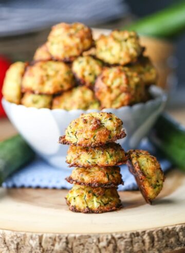 Cheesy zucchini bites stacked on a cutting board, with a bowl full of bites in the background.