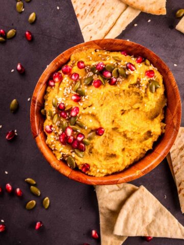 Bowl of pumpkin hummus topped with pomegranate arils and pumpkin seeds.