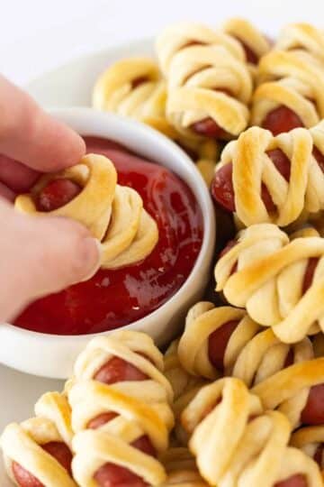 Mini hot dogs wrapped in crescent roll dough to look like mummies, with a hand dipping one into a red sauce.